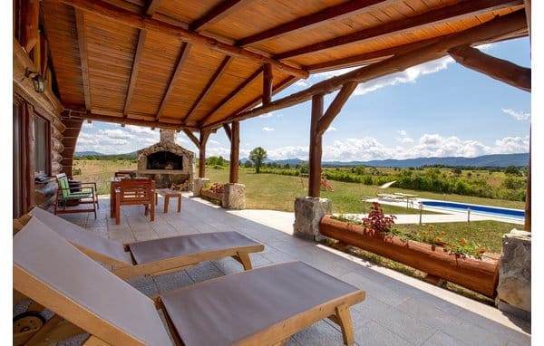 A spacious outdoor terrace features wooden beams and a tiled floor. Two lounge chairs are positioned under the roof, facing scenic views of rolling hills. An outdoor fireplace is present, with a glimpse of a swimming pool and lush greenery in the background.