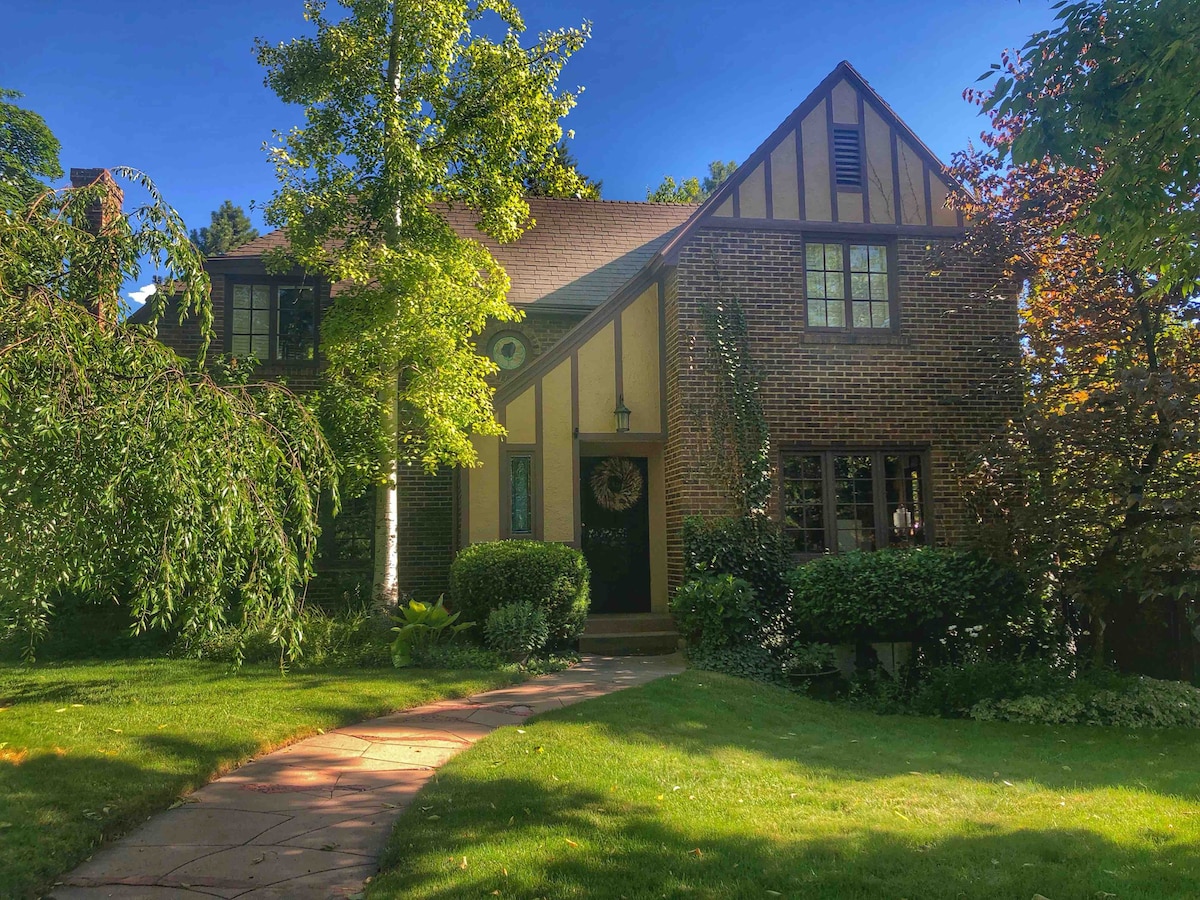 The exterior of a charming two-story brick house is visible, featuring a distinctive Tudor style with yellow accents. A well-maintained lawn highlights a stone pathway leading to the entrance, surrounded by lush greenery and colorful foliage from nearby trees.
