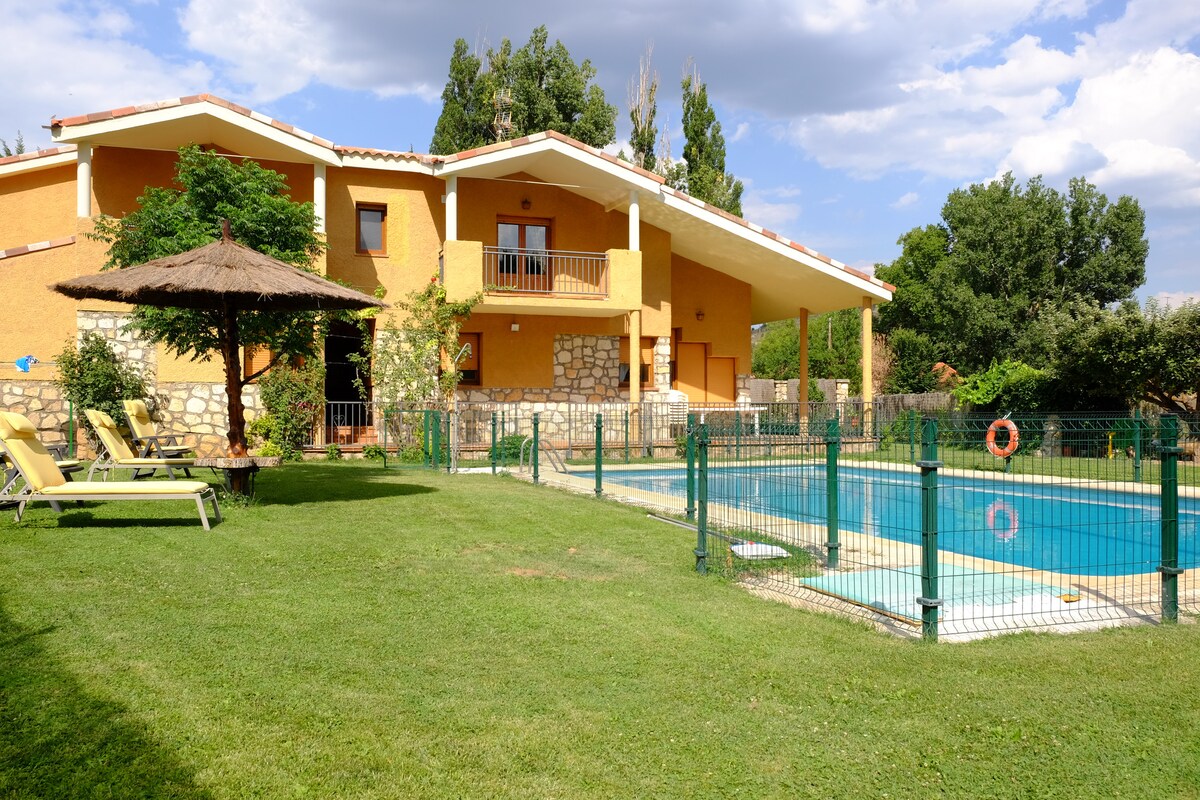 A spacious house features a large pool enclosed by a green fence. Sun loungers are arranged on one side of the pool, while lush trees and shrubs provide a natural backdrop. The two-story structure showcases a balcony and a thatched-roof hut for relaxing outdoors.