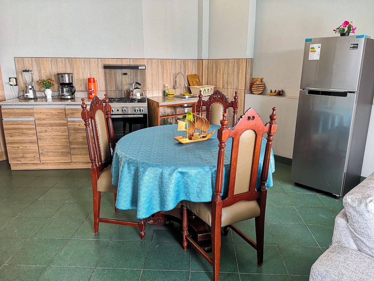 The kitchen area features a round dining table with ornate wooden chairs and a turquoise tablecloth. A well-equipped kitchen is visible, including a stove, refrigerator, and cabinets. Natural light illuminates the space, highlighting the modern appliances and countertops.