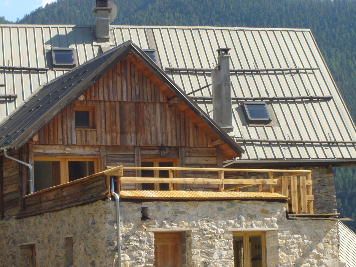 The exterior of the building features a blend of rustic wooden and stone elements, complemented by a sloped metal roof. A private terrace made of wood extends from the upper level, offering access to outdoor views. Skylights are visible on the roof.