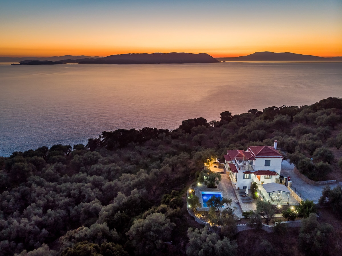 An aerial view captures the villa nestled among greenery, with a serene pool and patio illuminated against the twilight. The tranquil sea reflects shades of orange and purple as the sun sets over distant islands.