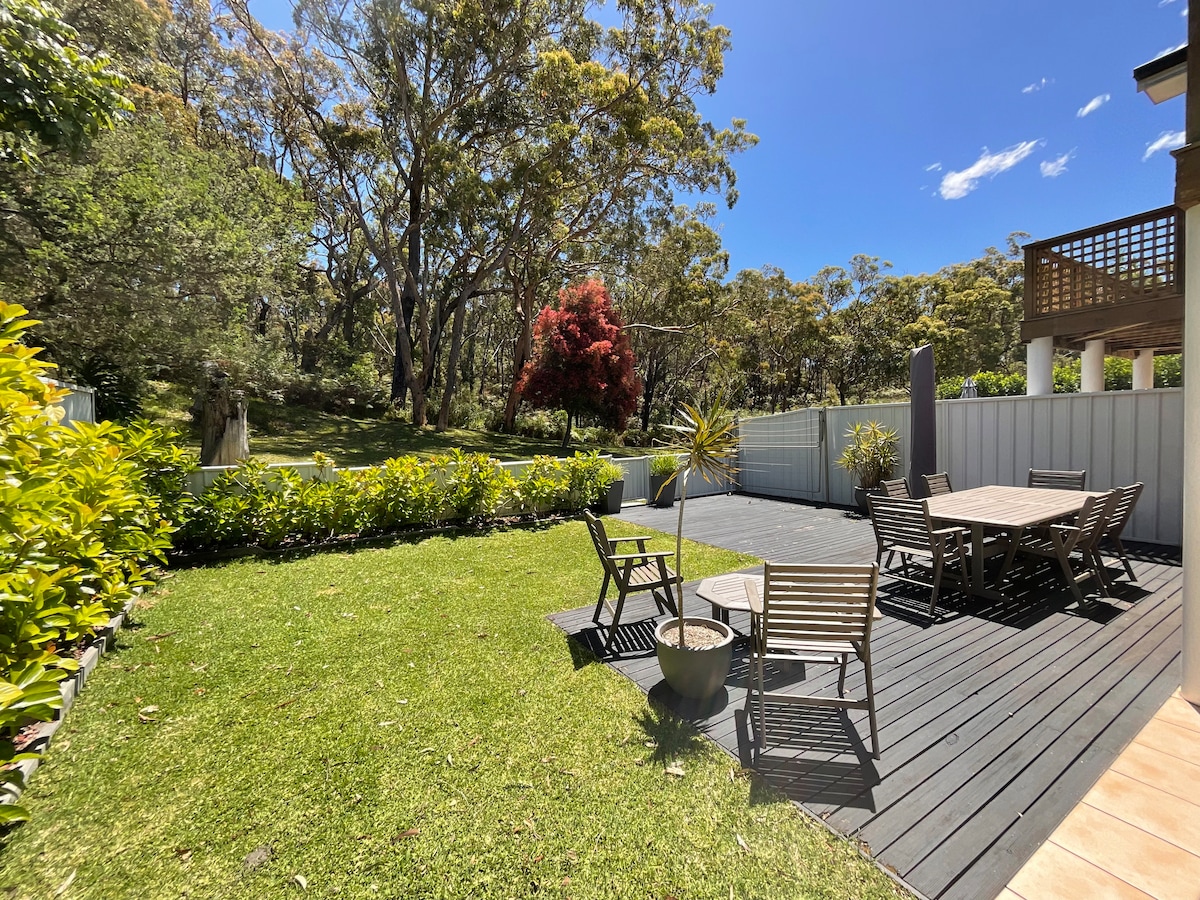 A spacious outdoor area features a wooden deck surrounded by lush greenery. Several seating arrangements are visible, including a large dining table and multiple chairs. Beyond the deck, landscaped gardens blend with the natural surroundings, providing a peaceful setting under a clear blue sky.