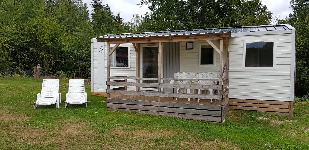 A mobile home features a spacious wooden terrace with a railing, accompanied by two white lounge chairs. The entrance is flanked by large glass doors offering natural light. Mature trees provide a green backdrop, enhancing the outdoor setting.