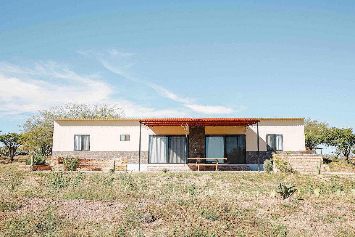 A modern single-story house is surrounded by open land under a clear sky. The façade features a mix of light-colored walls and a stone accent wall. Large glass doors provide access to a covered porch with a wooden bench, framed by low shrubs and desert plants.