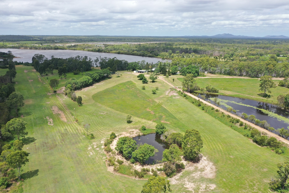 Aerial view of a sprawling green landscape with patches of forest and water bodies. A large open field is visible, bordered by trees, with a small pond at the center. The surrounding area shows a mix of greenery and agricultural land.