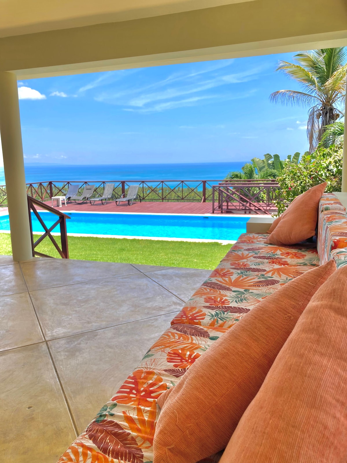 A shaded outdoor lounge area features a colorful couch adorned with tropical-patterned cushions. In the background, the private pool glistens under the sun, framed by lush greenery and offering panoramic ocean views that stretch to the horizon.
