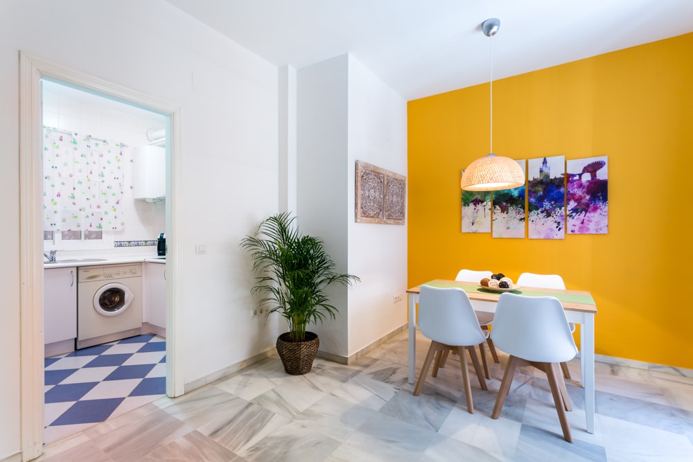 A bright dining area features a white table with four light wooden chairs, complemented by a colorful artwork on the wall. A potted plant adds a touch of greenery, while an adjoining kitchen is visible in the background, showcasing practical appliances.