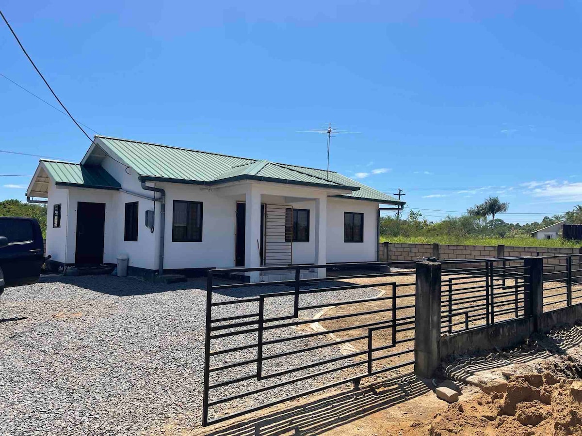 A modern house is presented against a clear blue sky. The exterior features a white facade with a green metal roof and multiple windows. A gravel driveway leads to the property, enclosed by a black gated fence. Green landscaping surrounds the area.