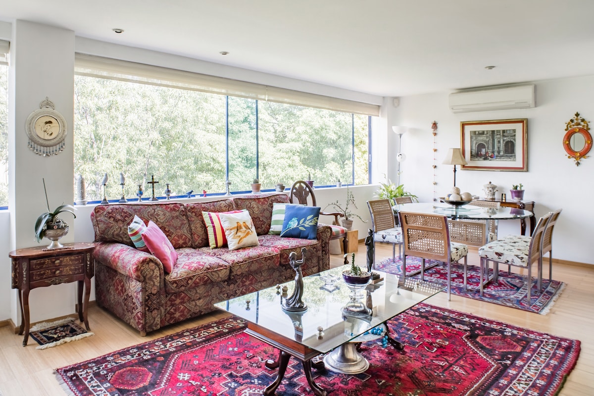 A spacious living area is filled with natural light, featuring a patterned sofa and wooden side table. A glass coffee table sits atop a decorative rug, while a dining area with woven chairs is positioned beside large windows. Green plants add a touch of nature.