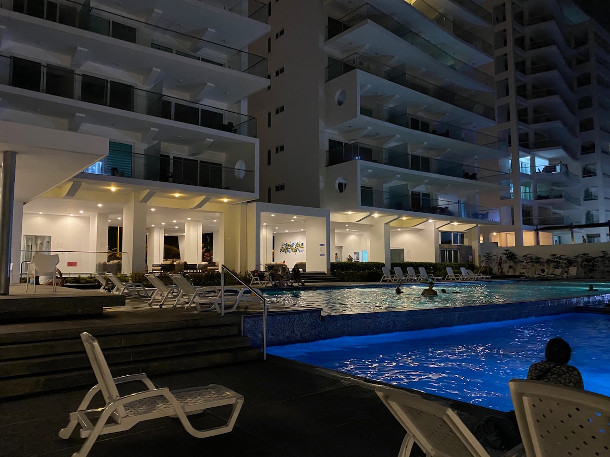 A well-lit pool area is shown during the evening, featuring a spacious pool with gentle illumination. Lounge chairs are arranged around the pool, while the building's balconies are visible in the background, reflecting a peaceful ambiance. Some guests are seen enjoying the water.