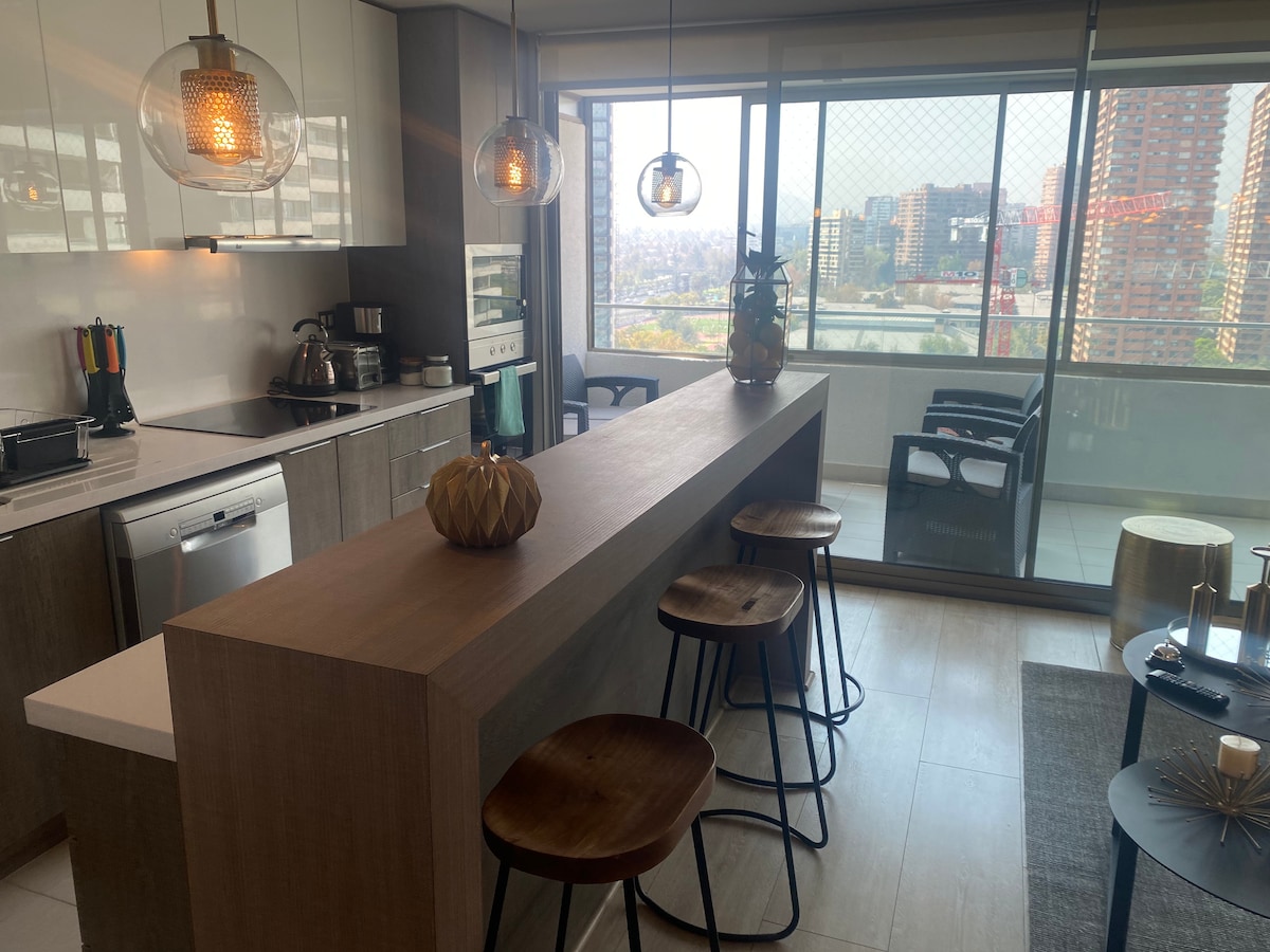 A modern kitchen is presented with wooden cabinetry and stainless steel appliances. A long countertop with three wooden stools faces a large window, offering a city view. Pendant lights hang above the countertop, creating a bright and welcoming atmosphere.