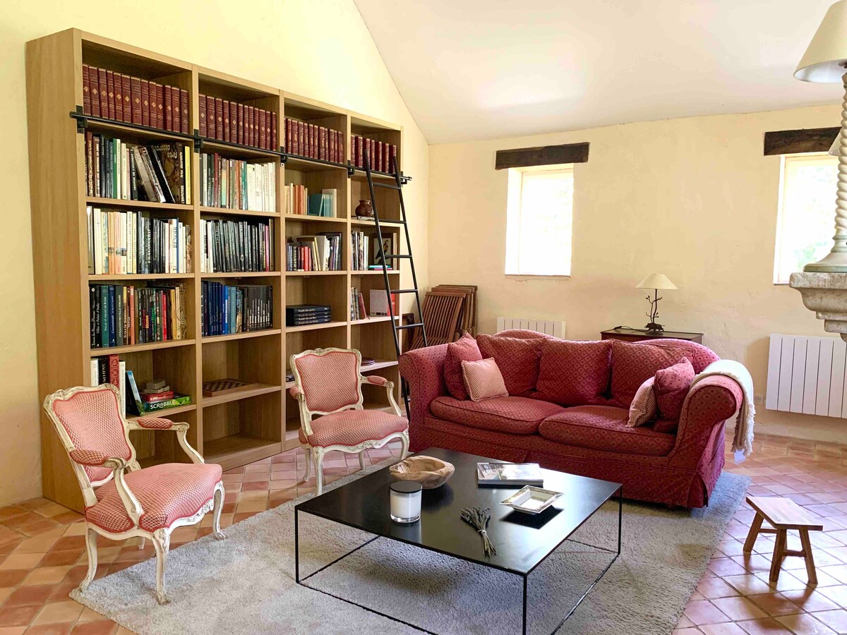 A spacious living area features a pink sofa and two elegant chairs facing a low coffee table. A tall bookshelf filled with books occupies one wall, and two windows allow natural light to brighten the room.