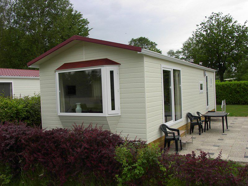 A modern chalet is shown, featuring light-colored wooden siding with a red roof. Large windows allow natural light to enter, and a small patio area with black chairs is visible. Lush greenery surrounds the chalet, enhancing the outdoor space.