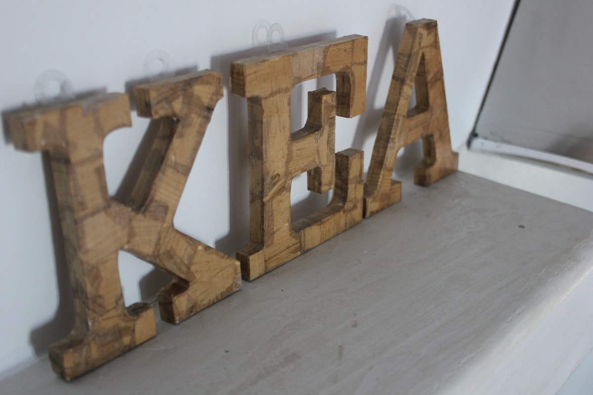 Wooden letters spelling 'KEA' are displayed on a smooth, light-colored surface. The letters have a natural finish, showcasing the grain and texture of the wood. A reflective surface is visible in the background.