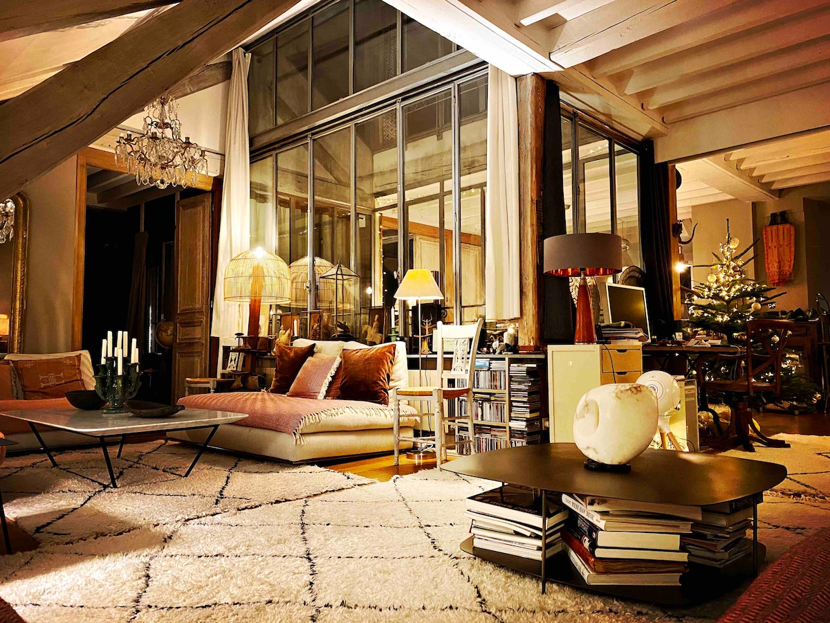 The spacious living area is characterized by high ceilings and large glass windows that allow natural light to fill the space. A large, comfortable seating area is complemented by a decorative coffee table stacked with books. Soft textiles and ambient lighting create a cozy atmosphere.