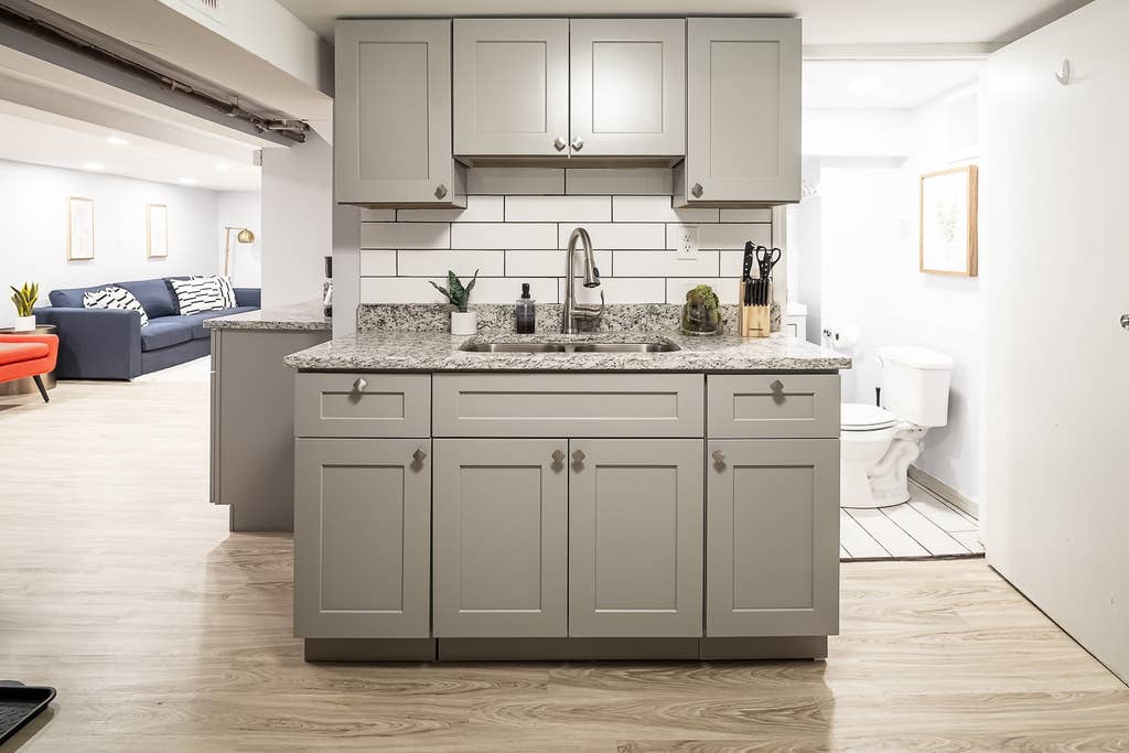 The kitchen area features modern gray cabinetry paired with granite countertops. A sink is positioned on the countertop, accompanied by kitchen utensils and a small plant. The open layout allows visibility of the living space in the background, enhancing a sense of connectivity.