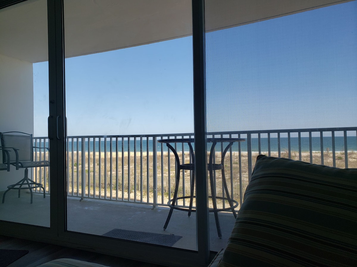 A wide glass door offers a view of the ocean and sandy beach beyond. Two outdoor chairs and a small table are positioned on the balcony, inviting relaxation. The clear blue sky enhances the coastal landscape seen from inside the living area.