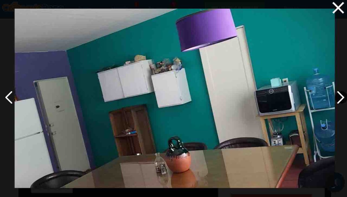 A dining area is depicted, featuring a large glass table surrounded by four black chairs. Bright turquoise walls complement a purple lampshade hanging overhead. Storage cabinets are mounted on the wall, and a water dispenser is positioned near the door, adding functionality to the space.