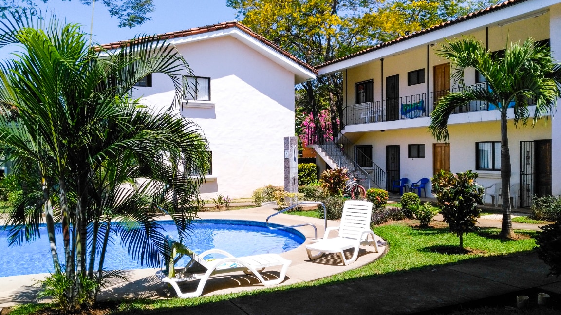 A well-maintained residential complex features a clear swimming pool surrounded by lush greenery. Sun loungers are positioned near the pool, and two buildings with balcony access are visible. Colorful landscaping adds to the inviting atmosphere of the outdoor space.