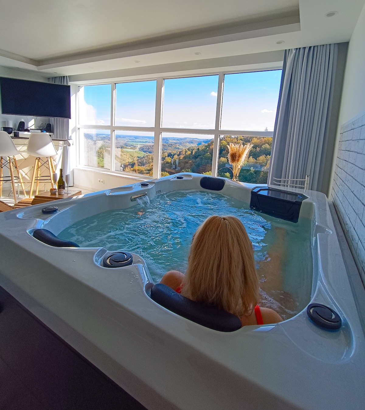 A spacious hot tub is positioned beside large windows offering a panoramic view of the landscape. Natural light fills the room, highlighting the water's surface. Modern furnishings, including high-top chairs, complement the serene setting, creating an inviting atmosphere for relaxation.