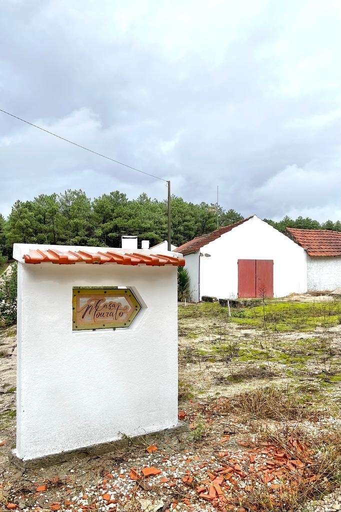 A charming white structure is visible, featuring a red-roofed sign that reads 'Casa do Monzato'. In the background, a series of white buildings with red doors blend into a natural landscape, framed by green trees and a cloudy sky.
