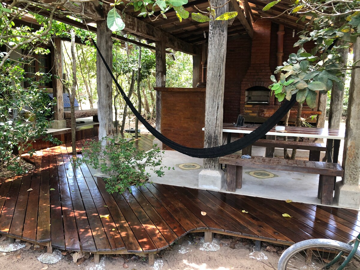 A shaded outdoor area features natural wooden decking and a spacious table with benches. A hammock is suspended between wooden beams, and a rustic barbecue area is visible in the background. Lush greenery surrounds the space, enhancing the connection to nature.