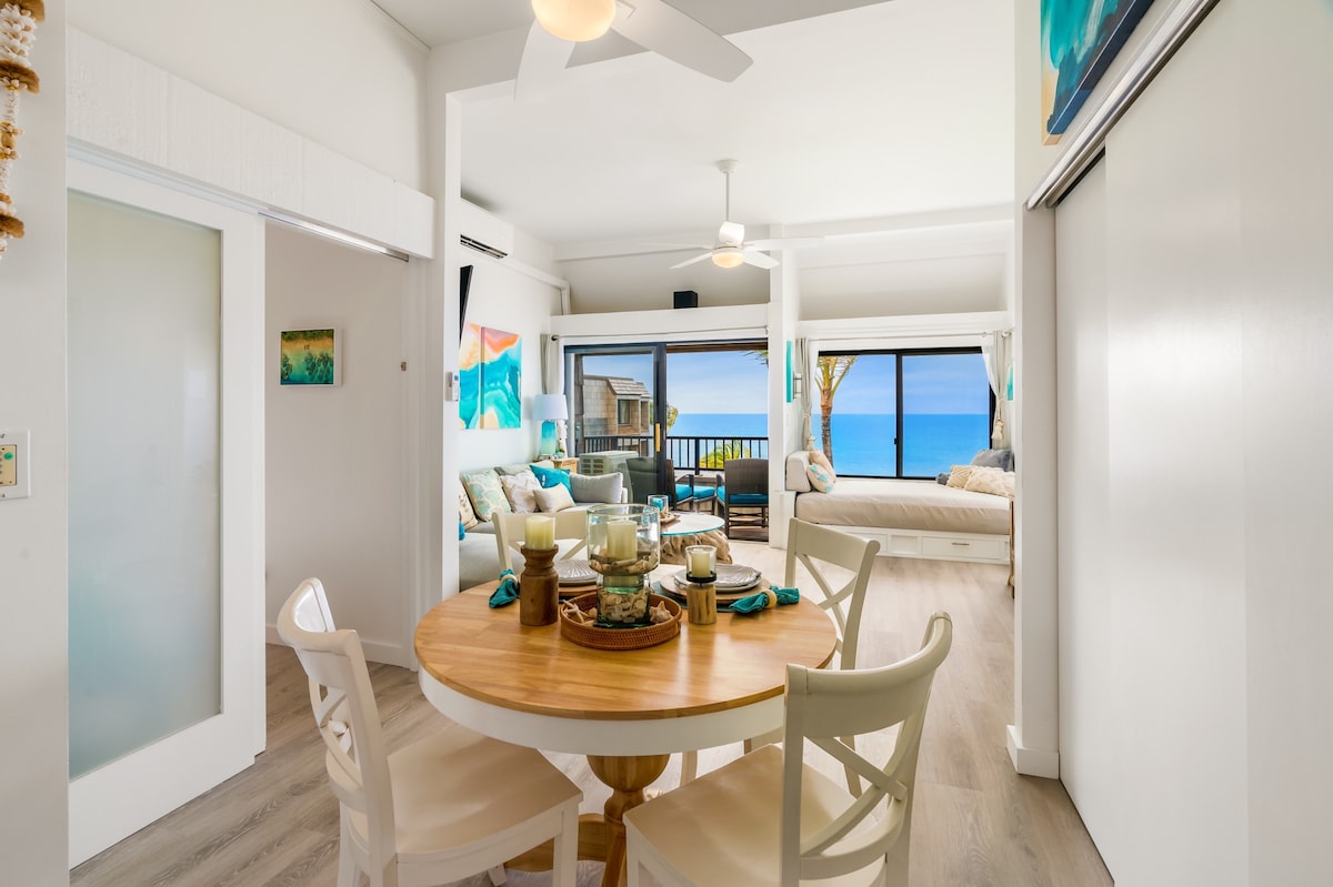A dining area features a round wooden table surrounded by white chairs, set with decorative items. The space is brightened by natural light coming through large windows that showcase panoramic ocean views. A cozy living area, complete with a daybed and colorful decor, is visible in the background.