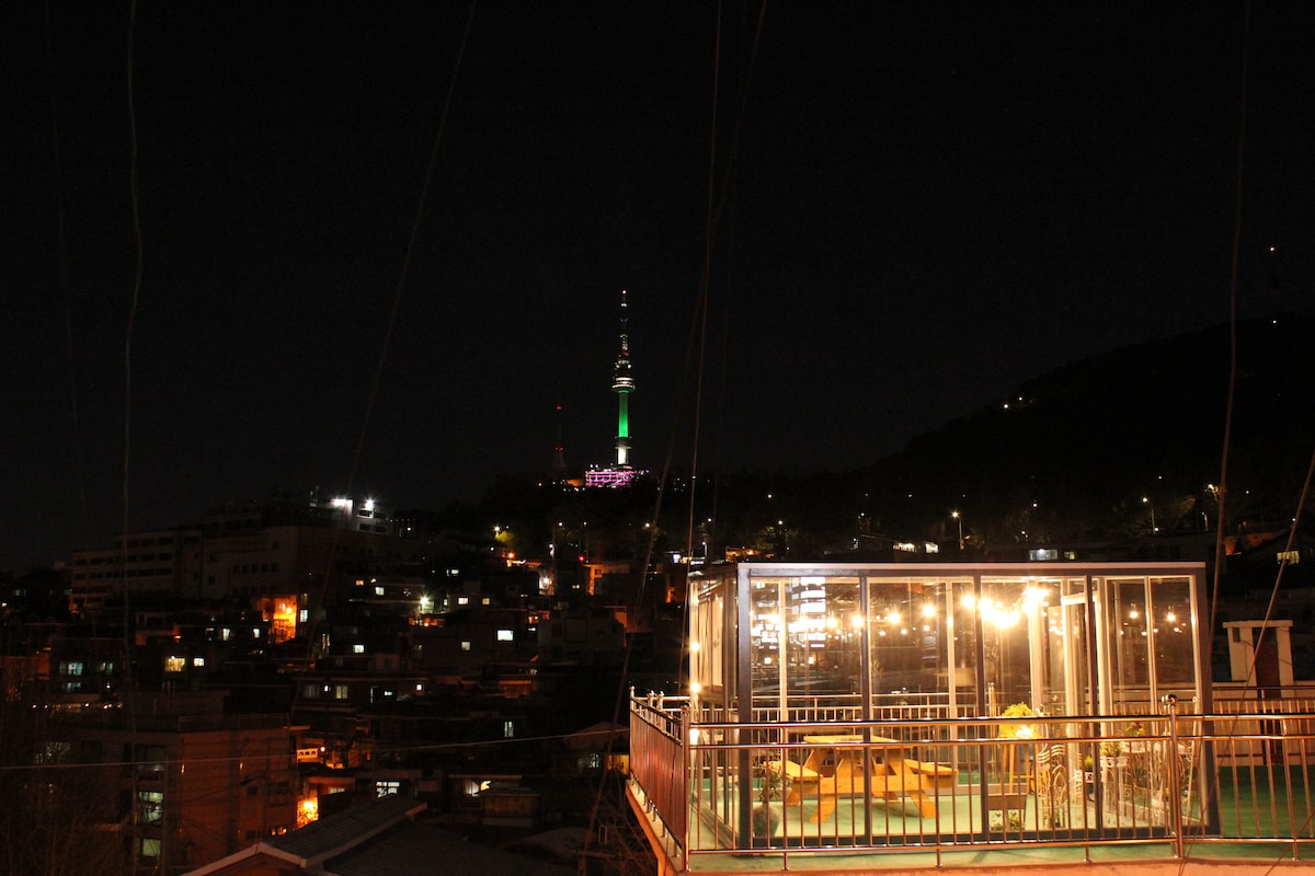 A rooftop area is showcased with a spacious seating arrangement, overlooking a vibrant cityscape at night. The iconic Namsan Tower is illuminated in colorful lights, providing a striking backdrop against the dark sky, while glowing city lights enhance the evening atmosphere.