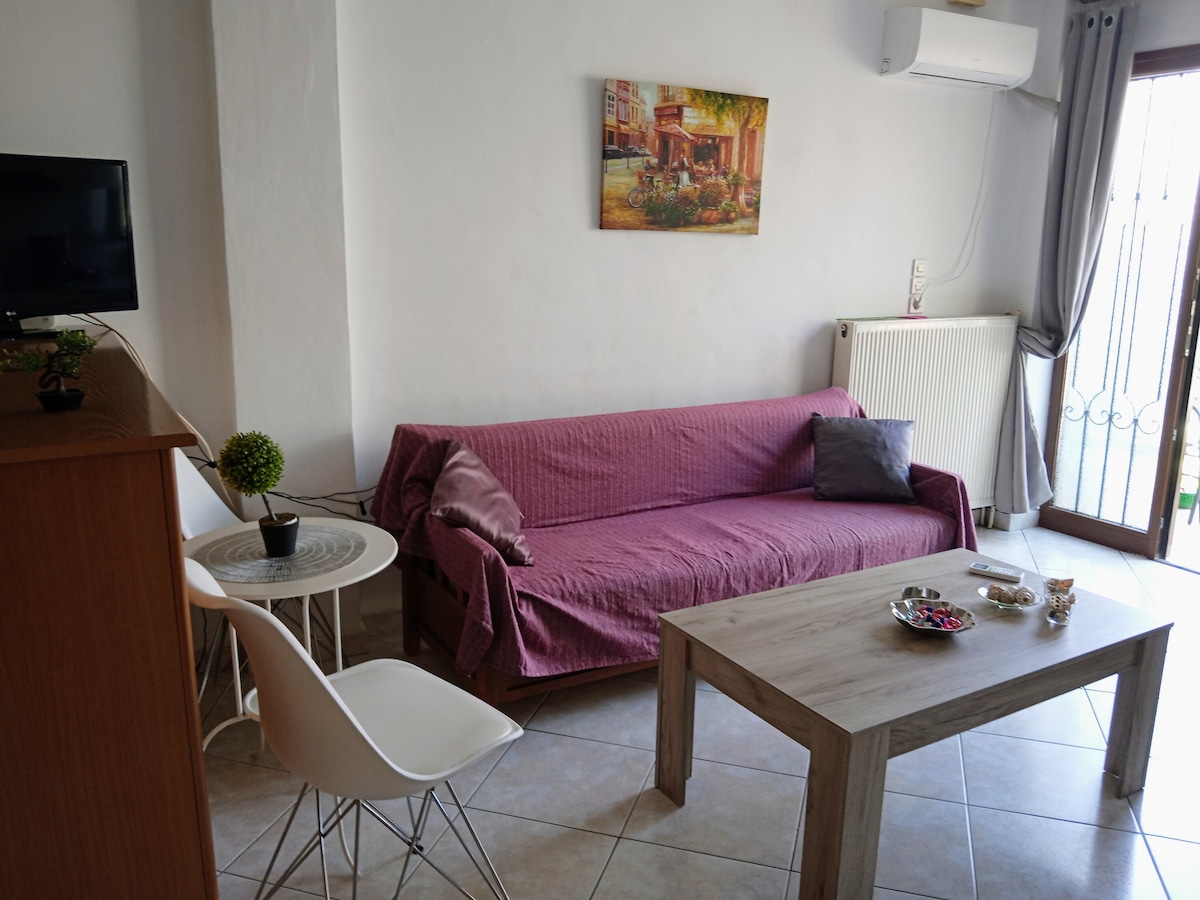 A welcoming living area features a purple sofa with cushions, complemented by a wooden coffee table. A small decorative plant sits on the table, and a wall-mounted TV is visible. Light enters through glass doors, with tasteful artwork hanging above the sofa.