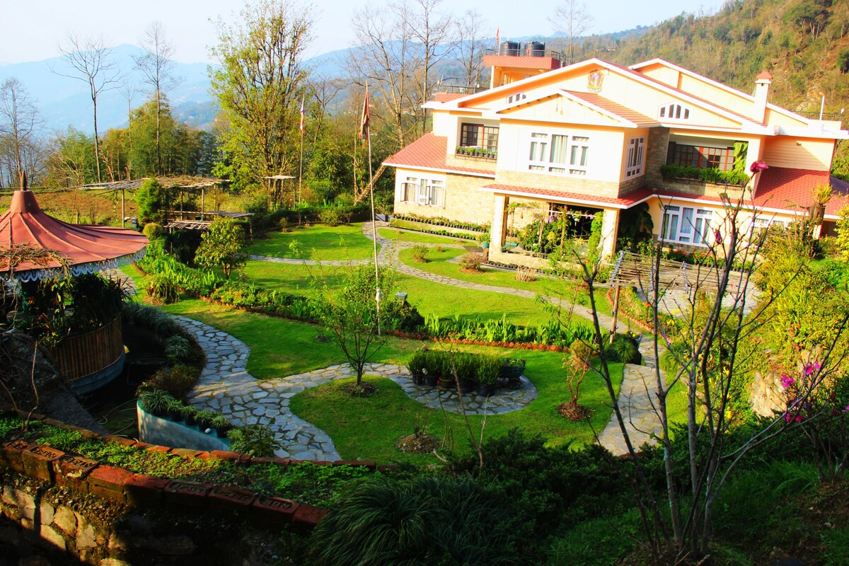 The homestay is set within a vibrant garden featuring neatly arranged stone pathways, lush greenery, and flowering plants. The modern two-story building boasts a red-tiled roof and large windows, surrounded by rolling hills and a serene landscape.
