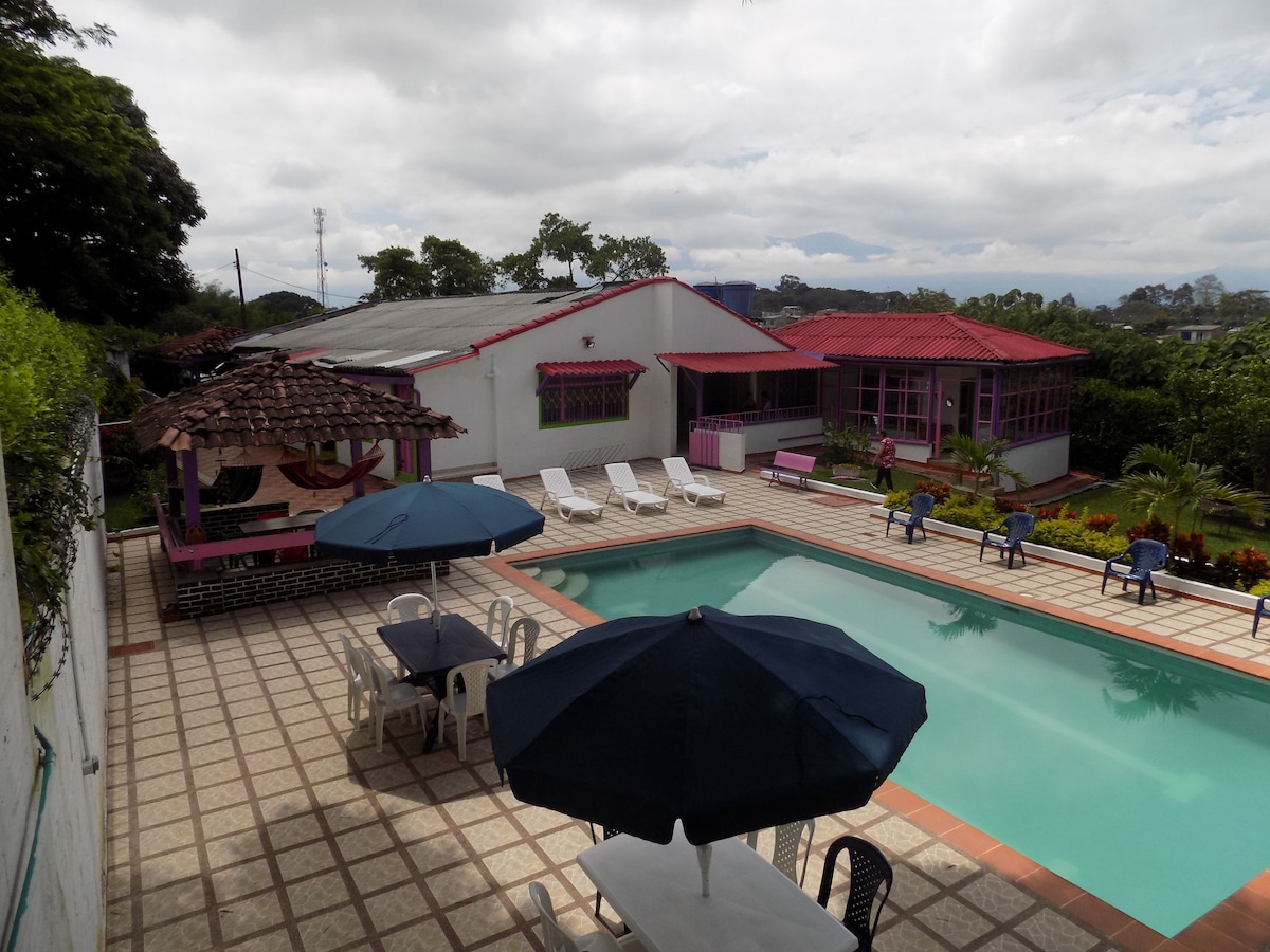 An outdoor area features a spacious swimming pool surrounded by lounge chairs. Umbrellas provide shade over tables set on a tiled surface. The vibrant architecture includes a bright red roof and large windows, complemented by lush greenery and flower beds in the background.