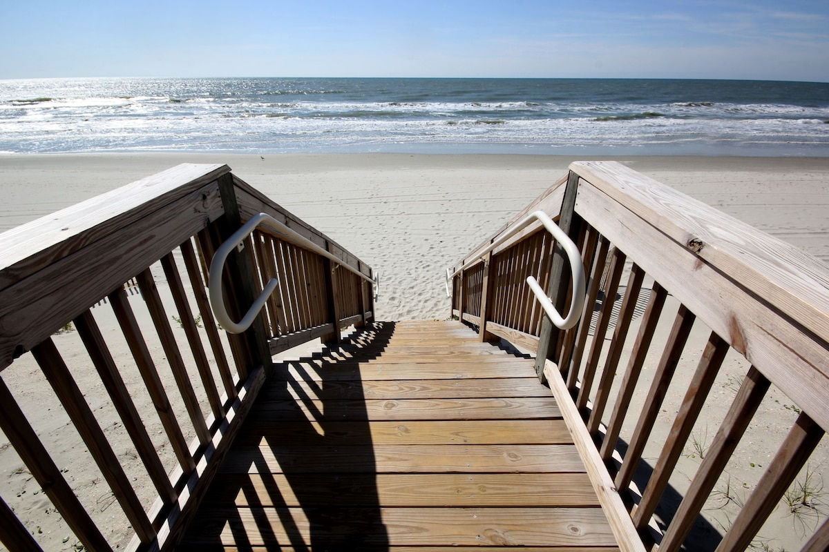 A wooden staircase leads down to a sandy beach, with sturdy handrails visible on either side. The calm ocean stretches out in the background, meeting a clear sky. Gentle waves can be observed lapping at the shore.