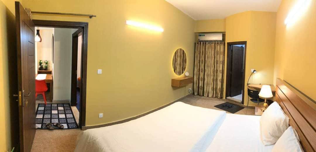 A warm-toned bedroom features a spacious bed with crisp white linens. A circular mirror is positioned above a wooden console, and a soft rug covers the floor. Nearby, a dressing area is visible, with patterned curtains framing the doorway.