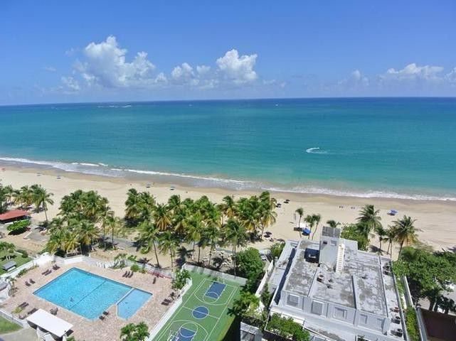 A panoramic view of the ocean and beach is presented, featuring soft golden sands and clear turquoise waters. A rectangular swimming pool is visible, alongside two tennis courts. Lush palm trees frame the scene, contributing to the tranquil coastal atmosphere.