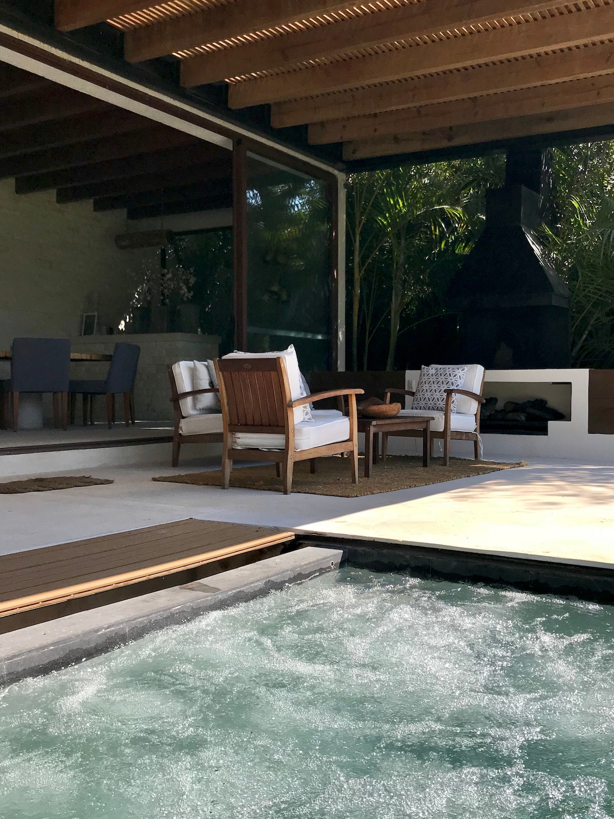 A relaxation area features two wooden chairs positioned on a patio beside a hot tub. The space is shaded by a slatted ceiling, and large glass doors provide a view into a dining area. Lush greenery is visible in the background, enhancing the setting.