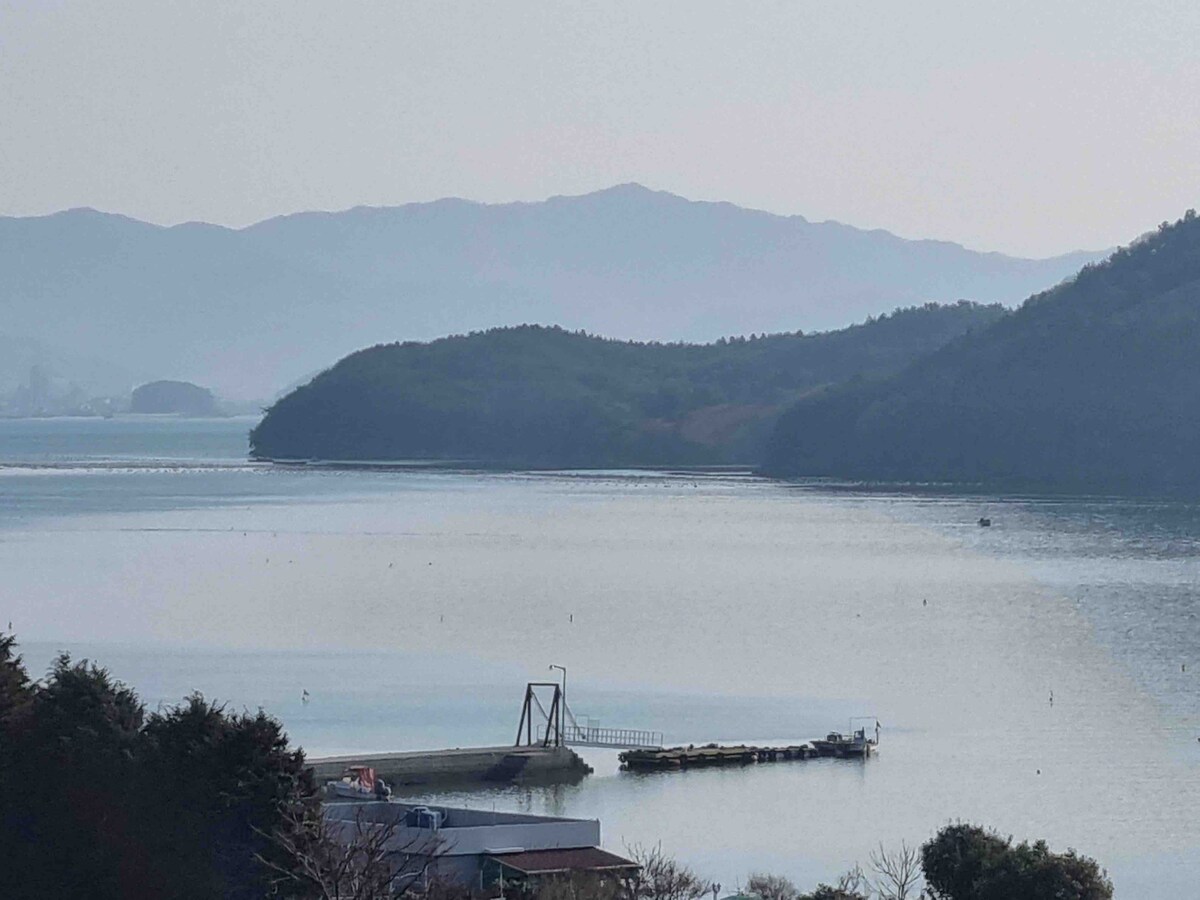 A serene view of a calm body of water, framed by rolling hills and distant mountains. A wooden pier extends into the water, with several boats anchored nearby. Soft reflections can be seen on the water's surface, creating a peaceful atmosphere.