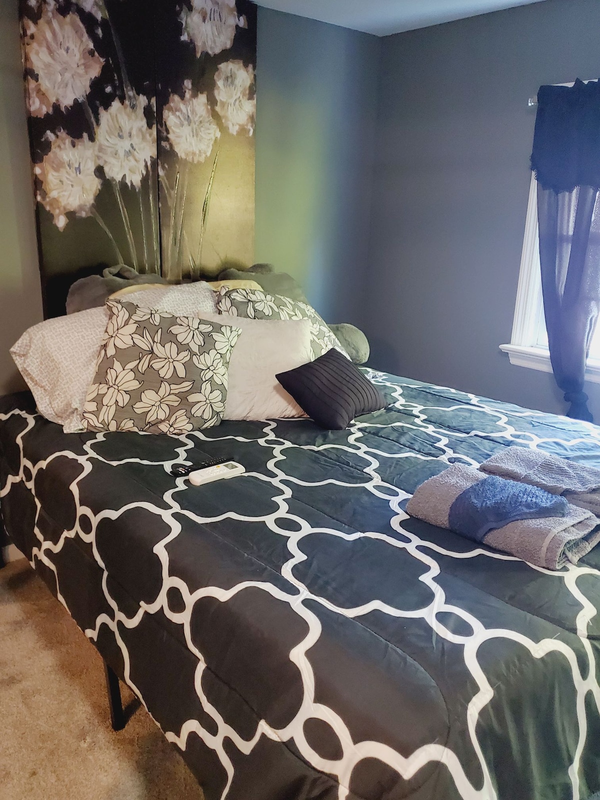 A cozy sleeping area features a patterned quilt on a spacious bed, adorned with various pillows. A floral artwork above the bed adds a decorative touch. Natural light enters through a nearby window, partially covered by dark curtains.