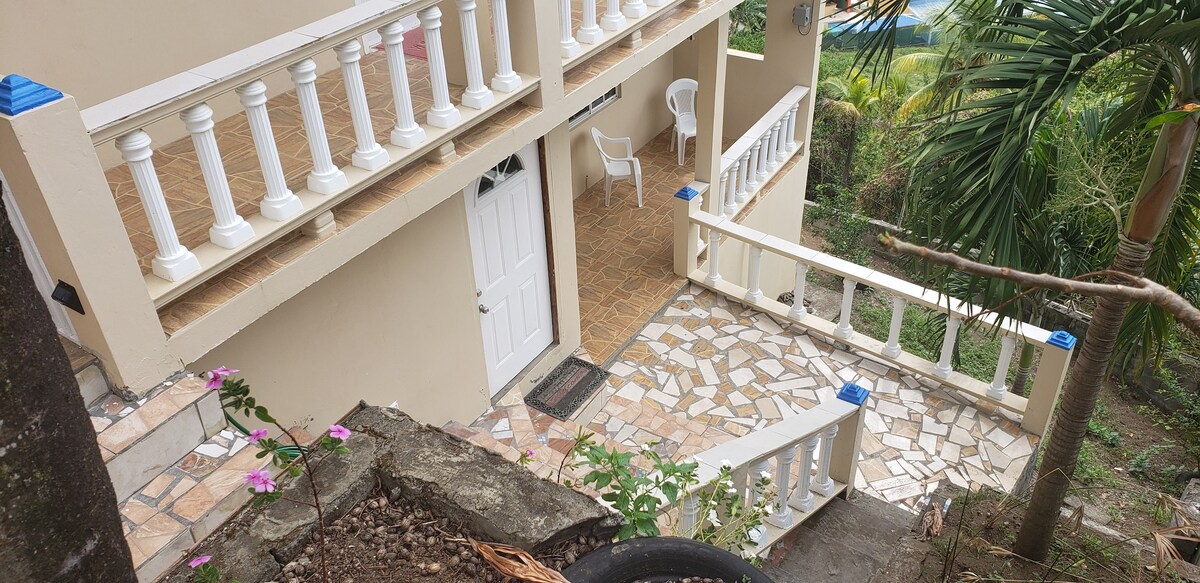 A view from a slightly elevated angle captures the exterior of a multi-level property featuring a tiled walkway. White railings border the stairs and upper balcony, while potted plants add greenery to the scene. Several white chairs are visible on the patio.