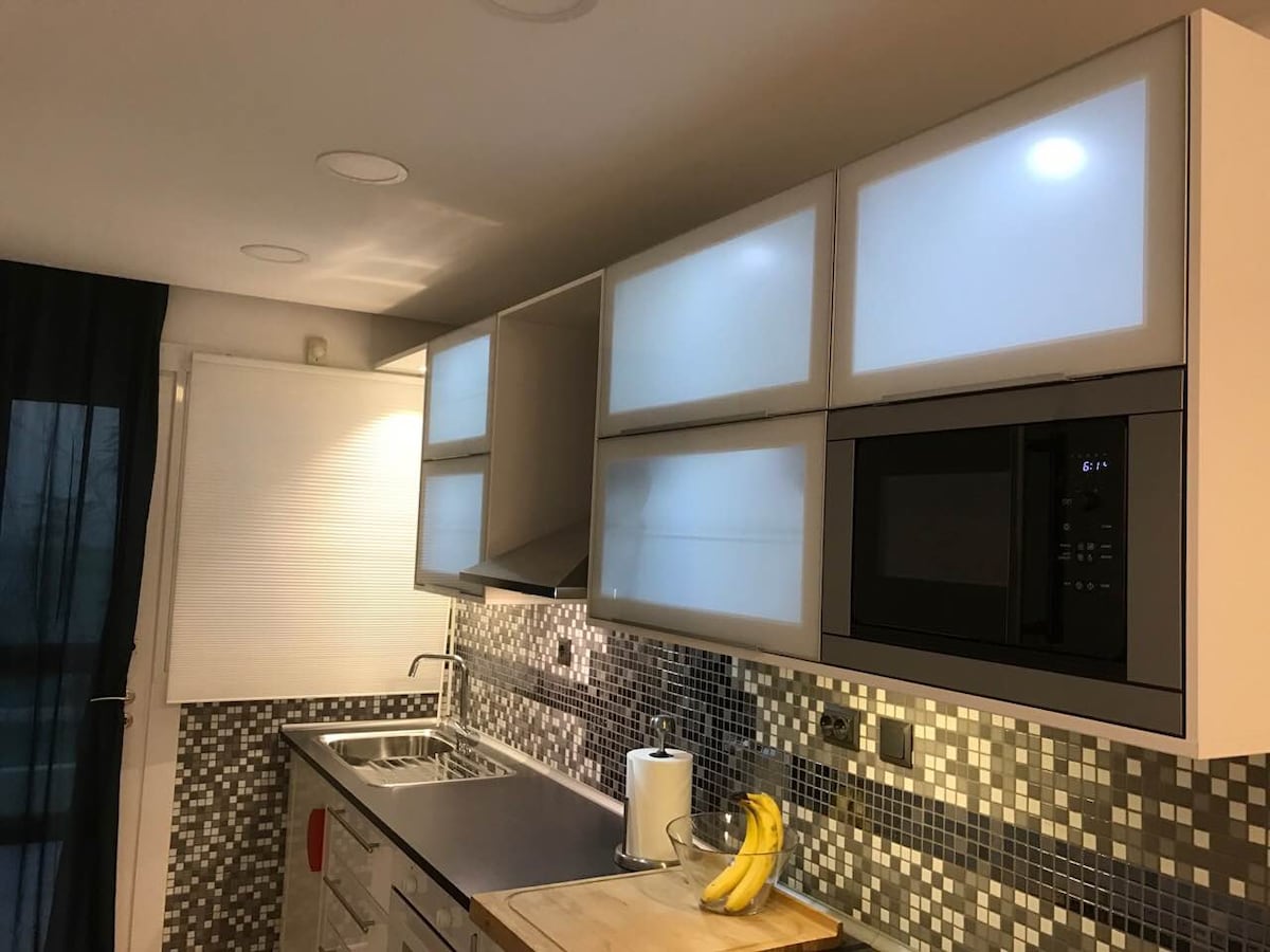 A modern kitchen features sleek cabinetry with frosted glass doors above a countertop. A stainless steel sink and a gas cooktop await use. The backsplash is adorned with mosaic tiles. Natural light enters through a window, complemented by stylish dark curtains.
