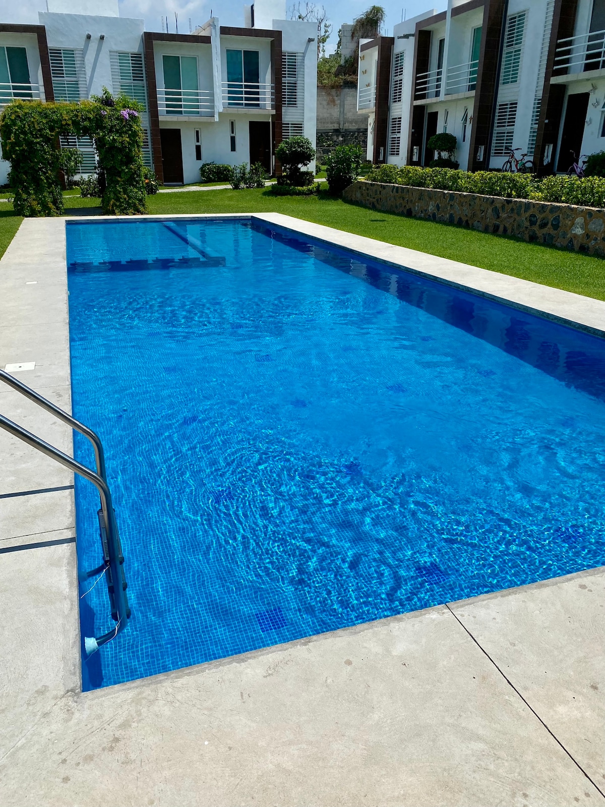 A large, inviting pool features clear blue water, surrounded by neatly manicured green lawns. Steps lead into the pool, and the serene environment includes nearby decorative plants and stones. The backdrop showcases modern residential structures.