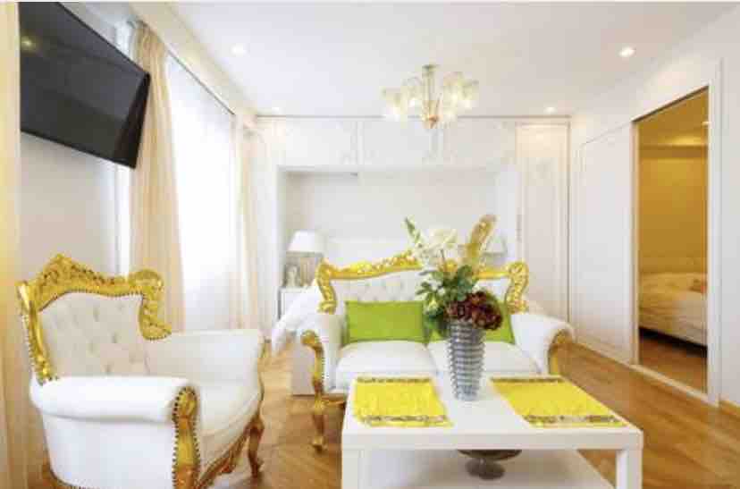 A bright and airy seating area features elegantly upholstered chairs with golden detailing and a white table at the center. A floral arrangement adds a touch of color, while natural light fills the space through large windows dressed with sheer curtains.