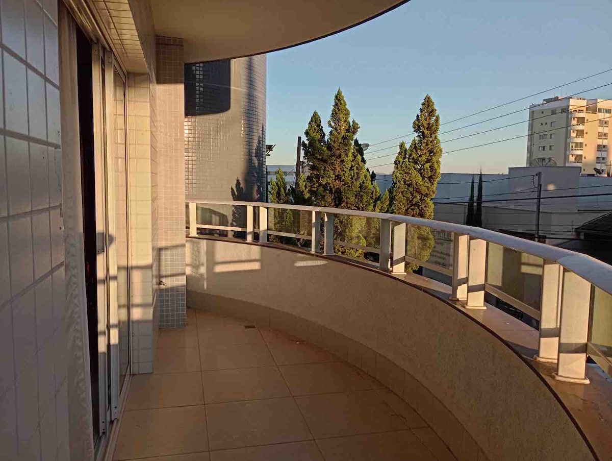 A curved balcony is shown with a smooth tile floor, surrounded by a low railing. Sunlight casts soft shadows on the surfaces, while views of nearby trees and buildings are visible in the background.