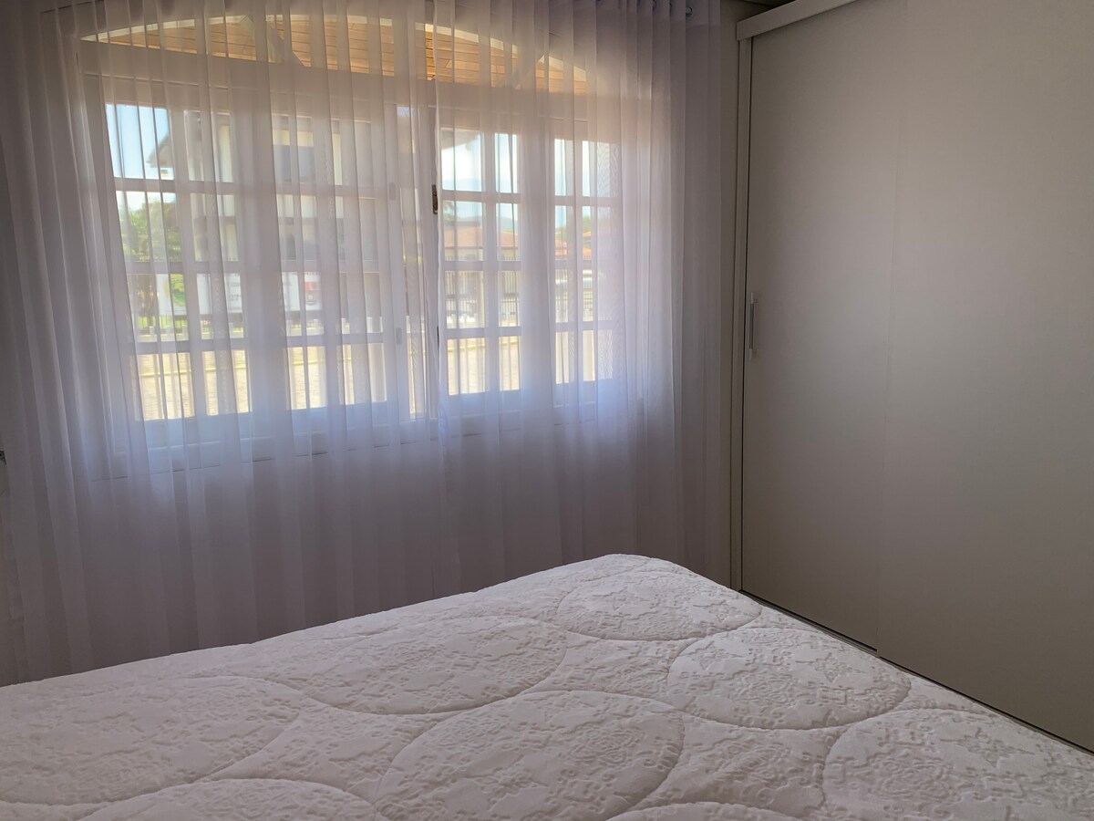 A bright bedroom features a spacious bed with a light-colored quilt. Sheer curtains allow natural light to enter, illuminating the room. A sliding door wardrobe is visible beside a window with multiple panes, providing a view of the outdoor area.