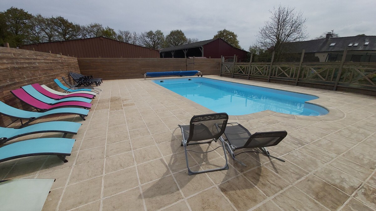 A spacious pool area features a rectangular swimming pool surrounded by a tiled deck. Colorful lounge chairs are arranged along one side, providing ample seating. A safety fence borders the area, and a few trees in the background enhance the natural setting.