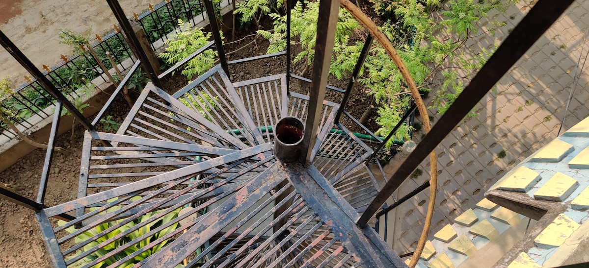 A spiral staircase made of metal descends from a terrace, surrounded by greenery. The design features alternating open spaces and horizontal bars, providing a view of the textured ground below. Sunlight softly illuminates the steps, creating a sense of calm in the shaded areas.