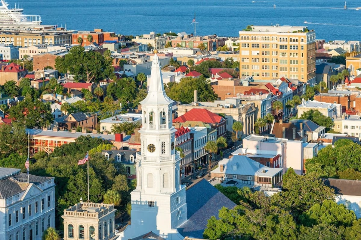 An aerial view captures the vibrant rooftops of downtown Charleston, featuring a mix of historic buildings with colorful facades and tree-lined streets. The distant coastline is visible, with boats dotting the harbor under a clear blue sky.