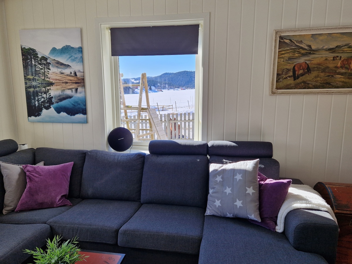 A spacious living area features a dark grey sectional sofa adorned with decorative pillows in shades of purple and white. A large window overlooks a scenic outdoor area, highlighting a wintry landscape. A piece of artwork depicting farm life is displayed on the wall.