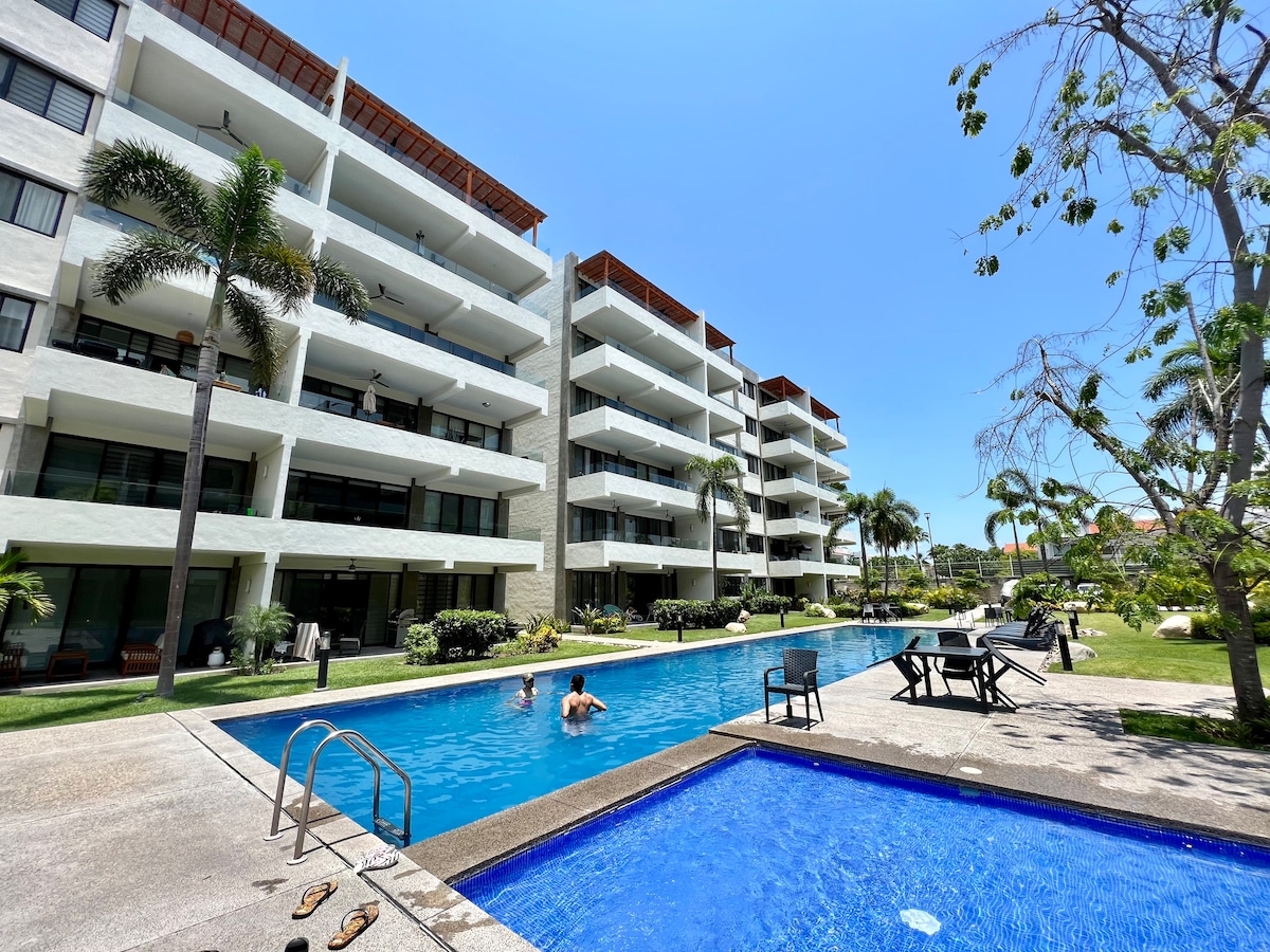 A tranquil pool area is surrounded by lush greenery and well-maintained landscaping. The three-story building features several balconies overlooking the pool, while lounge chairs and tables are positioned nearby for relaxation. Bright sunlight reflects off the clear water, creating a refreshing ambiance.
