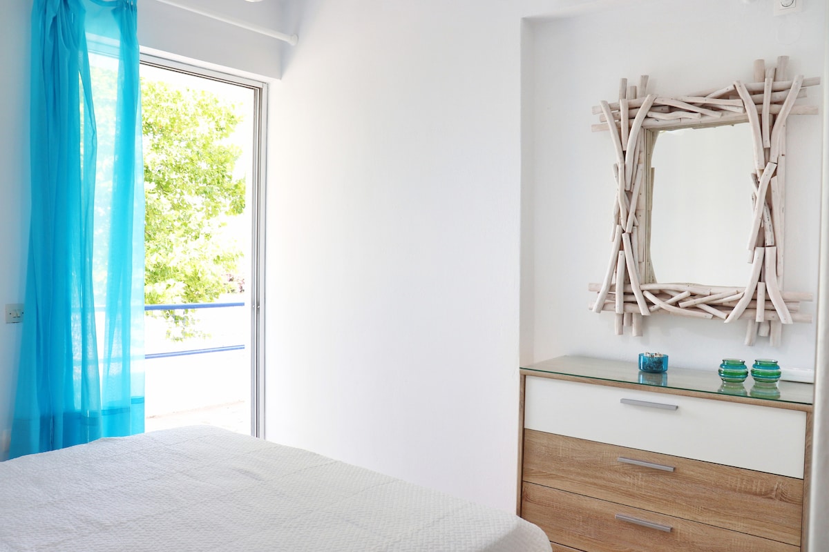 A bright bedroom features light-colored walls and a spacious bed covered with a quilt. Natural light enters through a large window adorned with sheer blue curtains. A decorative mirror framed in driftwood complements a modern dresser, enhancing the room's calming and airy ambiance.